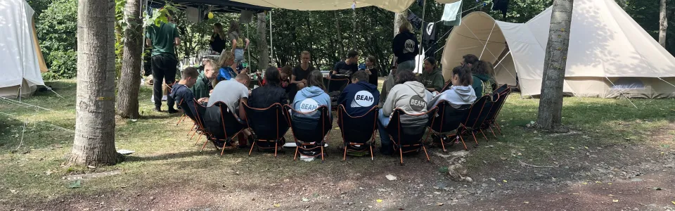 Deelnemers zitten onder een luifel bij de tenten